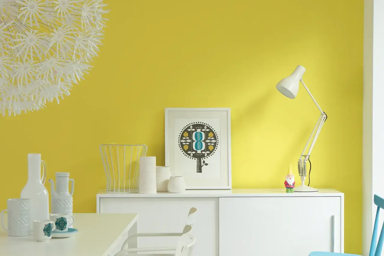 Parete giallo dorato illumina un mobile bianco con vasi, una lampada e un quadro. Un piccolo gnomo sorveglia la scena.