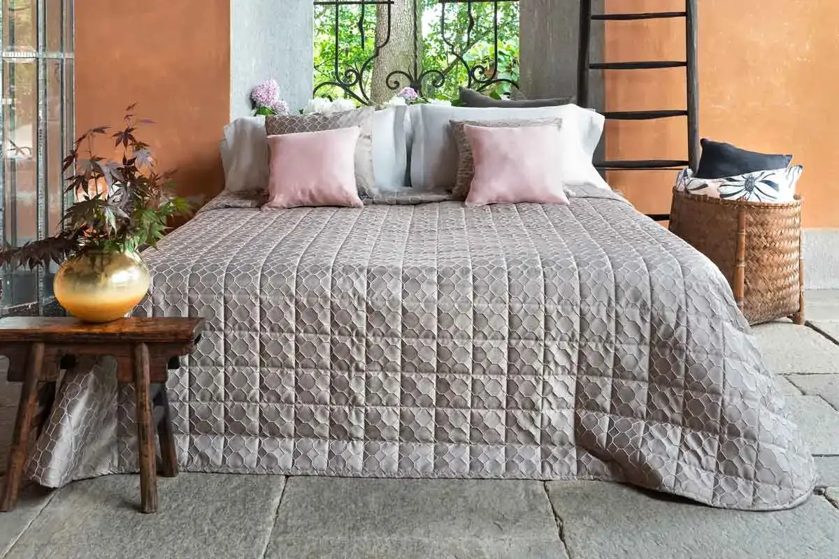 Letto con copriletto trapuntato grigio, cuscini rosa e vaso dorato. Ideale per lavare copriletto in lavatrice.