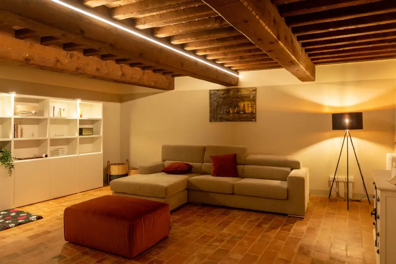 Illuminazione sopra divano crea un'atmosfera calda. Soggiorno con travi a vista, libreria e pouf rosso.