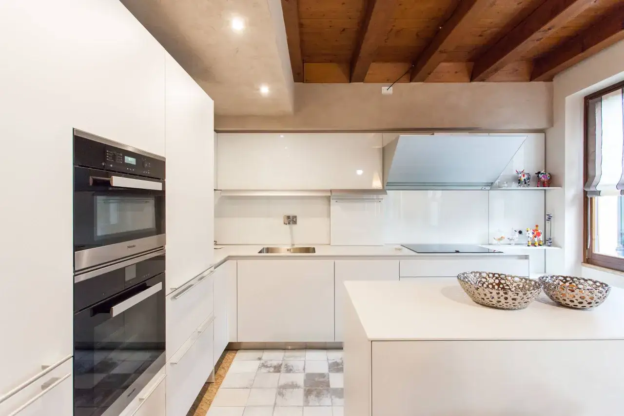 Cucina moderna con isola centrale, pensili bianchi lucidi e soffitto a vista con travi in legno. Ottime idee cartongesso soffitto cucina per illuminazione integrata.