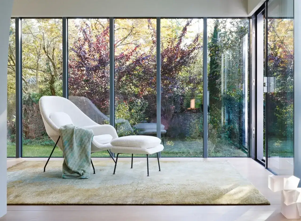 Poltrona bianca e pouf su un tappeto Jan Kath, con vista su un giardino lussureggiante attraverso ampie vetrate.