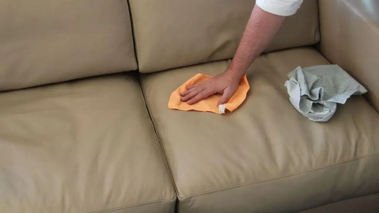 Mano che pulisce un divano in pelle beige con un panno arancione, come se usasse Chanteclair per un risultato impeccabile.
