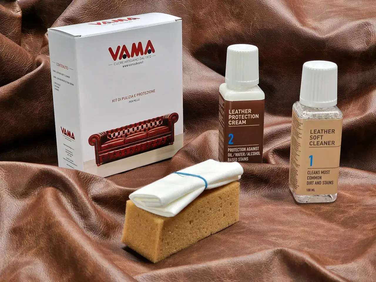 Kit Vama per la cura della pelle anilina: pulitore, crema protettiva, spugna e panno su sfondo in pelle marrone.