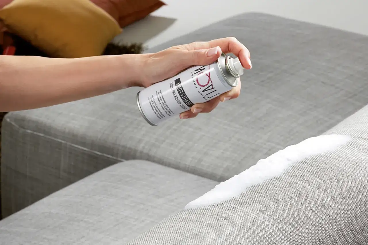 Mano che spruzza un prodotto su un divano grigio, un modo efficace per come togliere macchie dal divano.