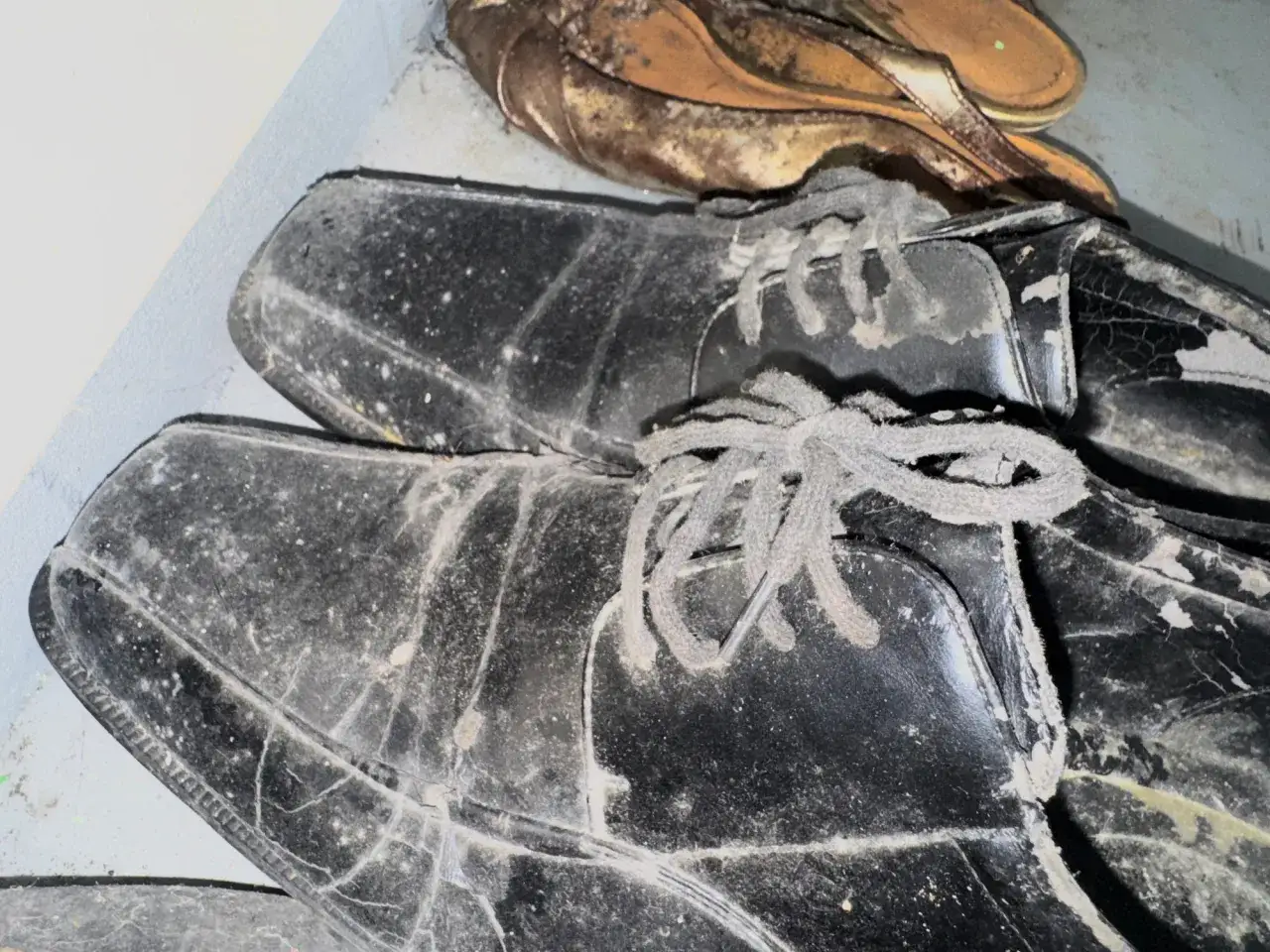 Scarpe nere e sandali dorati coperti di polvere e muffa su tessuti, abbandonati in un angolo.