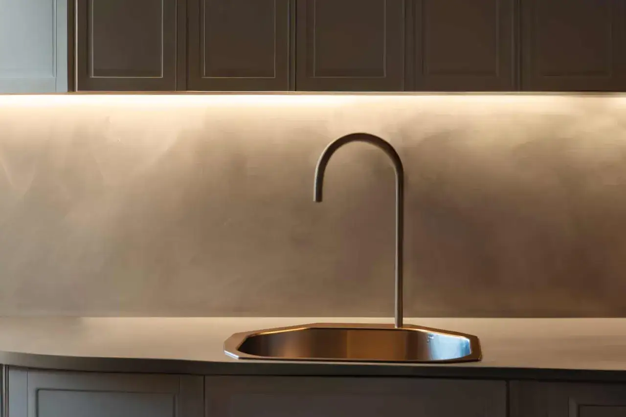 Rubinetto moderno su lavello in metallo, con parete di fondo in resina dietro cucina illuminata da una luce led.