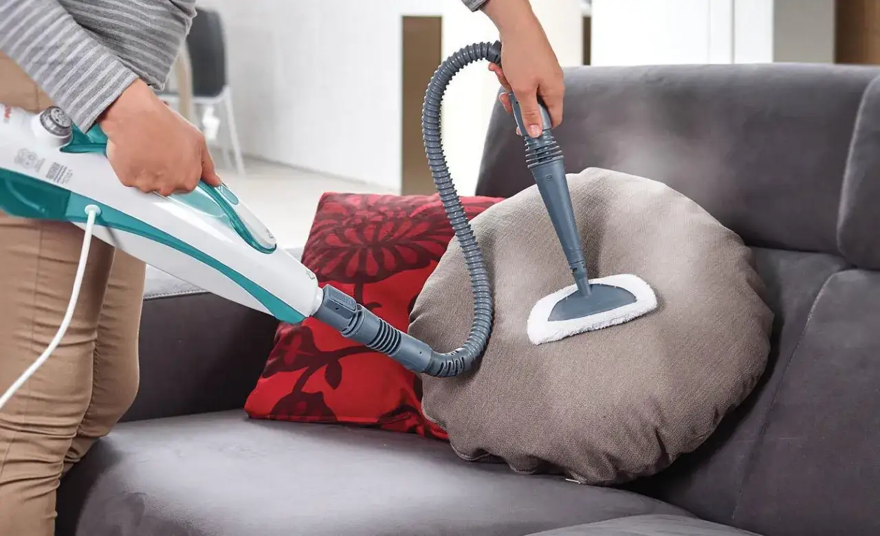 Persona pulisce un cuscino del divano in tessuto con un pulitore a vapore, mostrando come pulire il divano in tessuto in modo efficace.