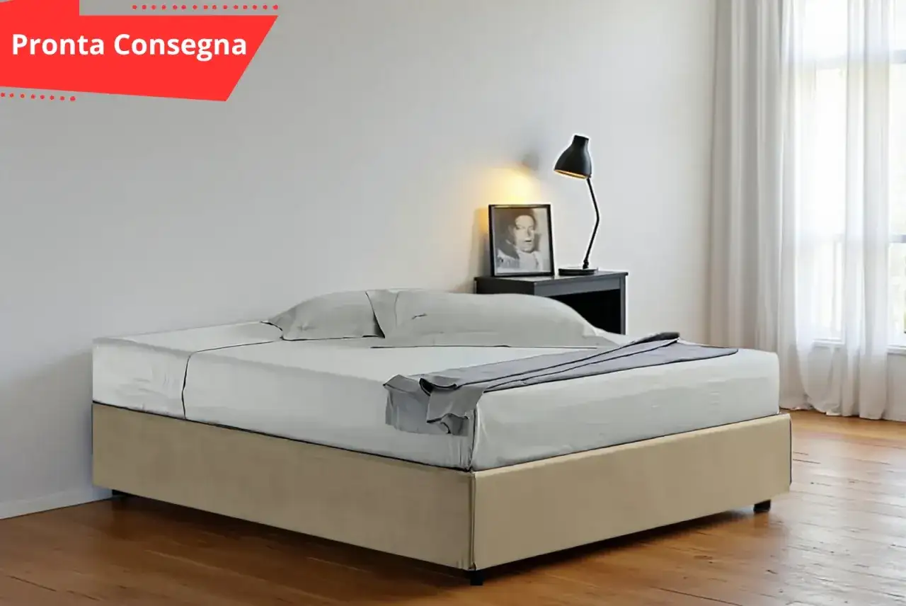 Un letto con sommier beige, lenzuola bianche e un plaid grigio. Sul comodino, una lampada e una foto.