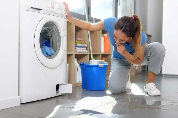 Donna preoccupata controlla la lavatrice che perde acqua da sotto. Sul pavimento bagnato, un secchio blu e uno straccio.