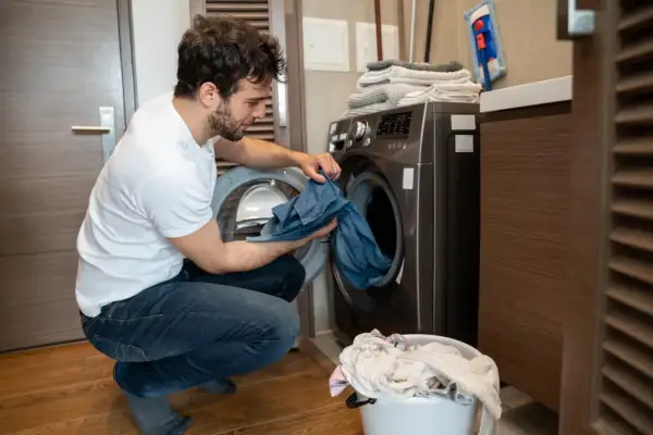 Uomo con maglietta bianca e jeans che carica la lavatrice, preoccupato per come eliminare le macchie da trasferimento di colore blu da un capo.