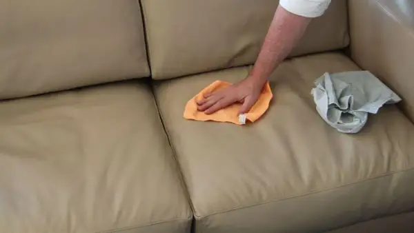 Mano che pulisce un divano in pelle beige con un panno arancione. Un altro panno grigio è appoggiato sul divano.