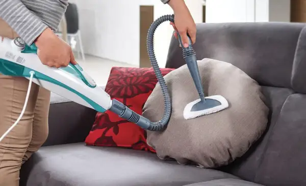 Come togliere macchie dal divano con un pulitore a vapore. La persona pulisce un cuscino del divano con un accessorio a spazzola.