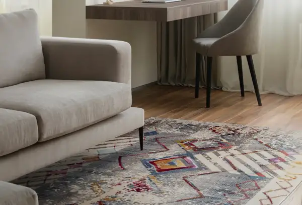 Un tappeto colorato sotto un divano beige, con una scrivania e una sedia sullo sfondo. Tappeto sotto divano si o no? La risposta è sì per aggiungere stile.