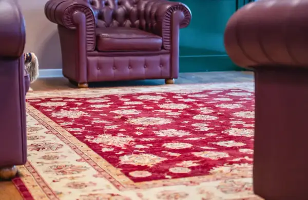 Un tappeto Yalameh rosso con motivi floreali chiari, adagiato su un pavimento in legno, accanto a poltrone in pelle bordeaux.