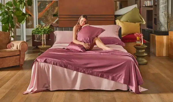 Una donna sorride su un letto con lenzuola rosa. La trapunta nuova va lavata prima di usarla.