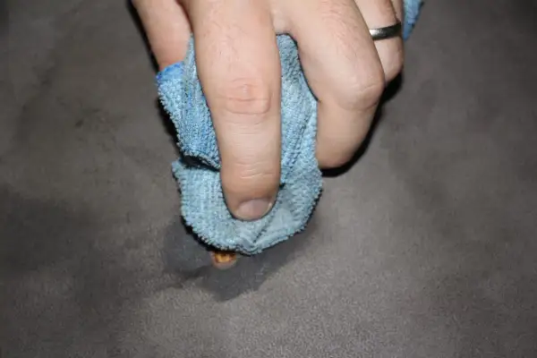 Mano che pulisce una bruciatura da sigaretta su tessuto grigio con un panno blu.