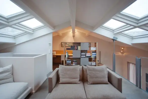 Accogliente salotto con soppalco, illuminato da lucernari. Divano beige, libreria moderna e scrivania in legno.