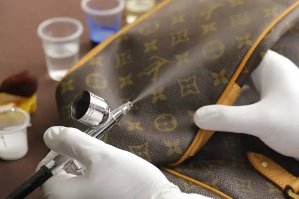 Mani guantate usano un aerografo per pulire la similpelle di una borsa monogrammata, mostrando un metodo per come pulire la similpelle.